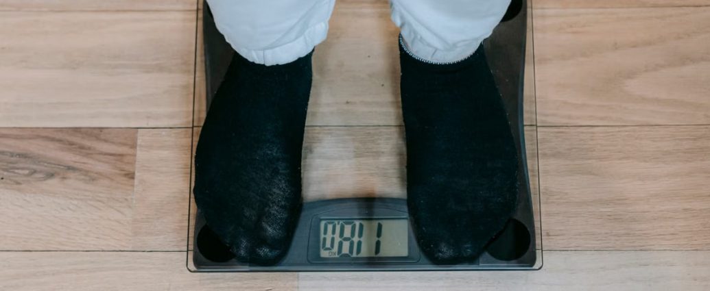 From above of crop unrecognizable plus size person in casual clothes standing on weighing scale on wooden floor