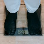 From above of crop unrecognizable plus size person in casual clothes standing on weighing scale on wooden floor