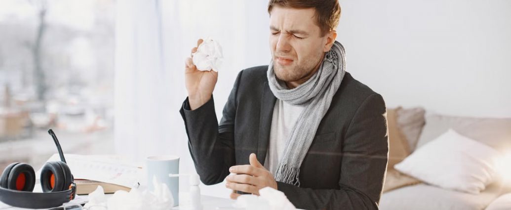 A sick man wearing a scarf sitting at his desk, holding a tissue, displaying signs of discomfort.