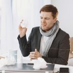 A sick man wearing a scarf sitting at his desk, holding a tissue, displaying signs of discomfort.