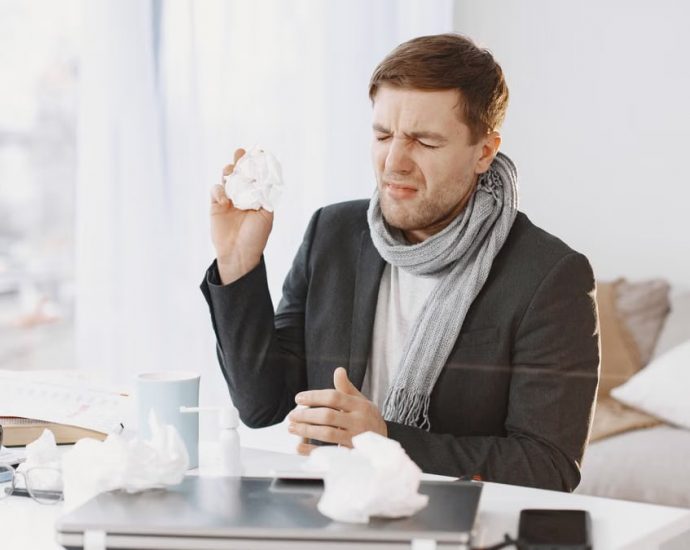 A sick man wearing a scarf sitting at his desk, holding a tissue, displaying signs of discomfort.